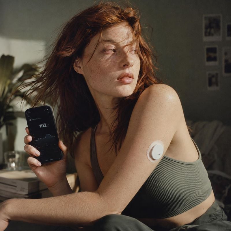 wearable glucose monitor and smartphone app equipment in use product photo: A red-haired woman in a ribbed sports bra holds up a phone displaying glucose data…