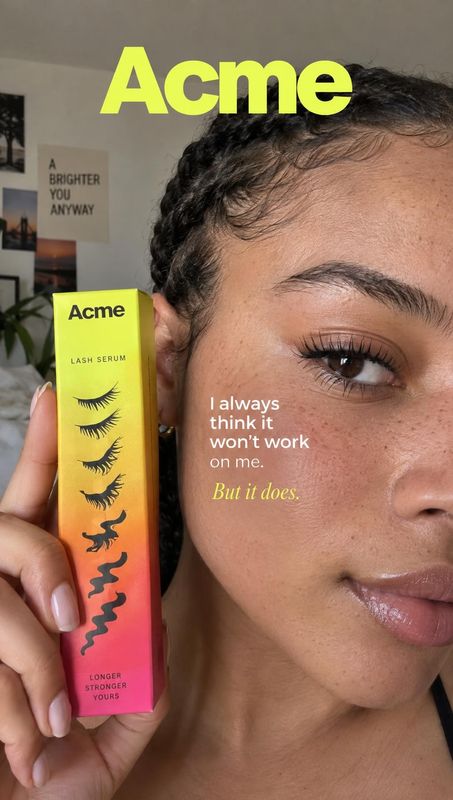 lash serum product in hand product photo: A woman holds a brightly colored lash serum box beside her face in a close-up interior portrait, with bold yellow bra…