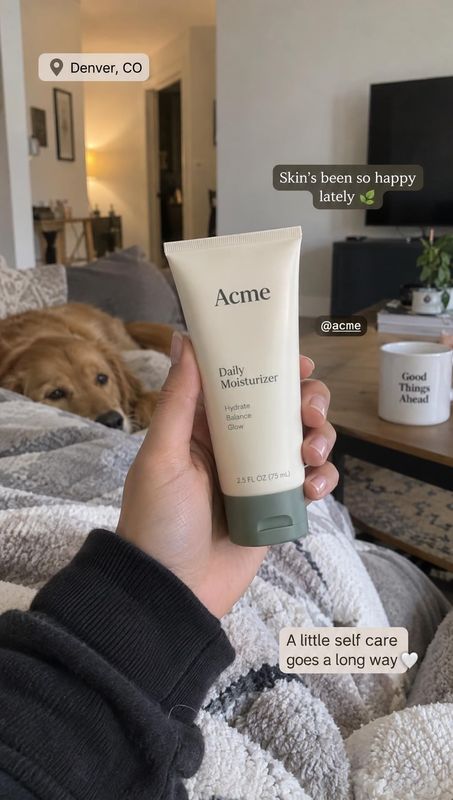 daily moisturizer product in hand product photo: A hand presents a tube of daily moisturizer in a cozy living room, with a dog resting on a blanket and warm, c…