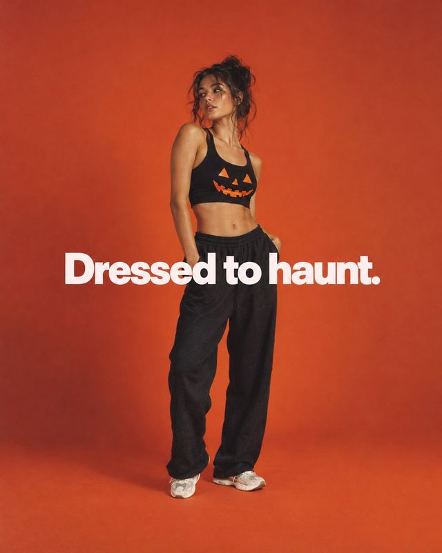 Halloween sportswear outfit halloween product photo: A model poses in a black pumpkin-print sports bra and loose trousers against a vivid orange studio backdro…
