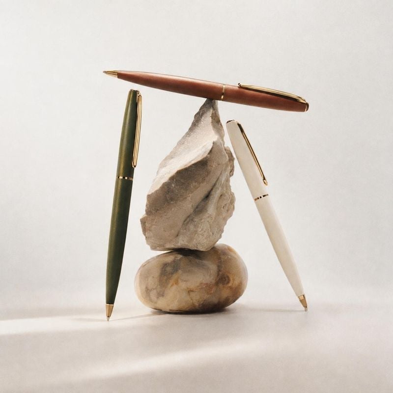 pens stationery product photo: Three elegant pens in muted green, ivory, and warm brown balance against sculptural stones on a softly lit neutral studio backdr…