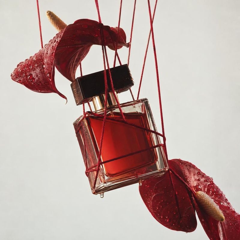 perfume floating product product photo: A red glass perfume bottle hangs among glossy, water-dappled crimson leaves against a pale studio background, creating…