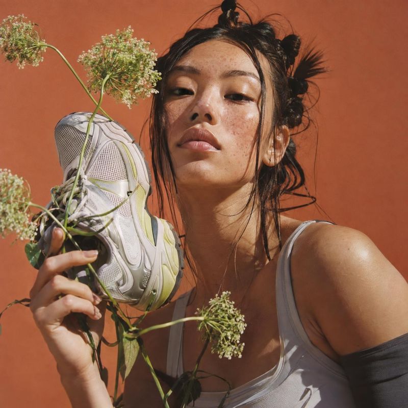 sneaker close-up detail product photo: A woman with loosely pinned hair holds a chunky white sneaker among delicate wildflowers against a warm terracotta studi…