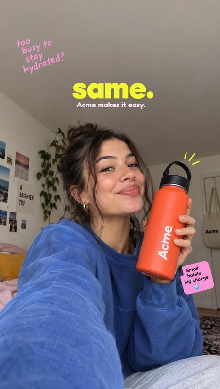 water bottle lifestyle scene product photo: A smiling woman in a blue sweatshirt holds a bright orange water bottle in a casual bedroom selfie, with playful pi…
