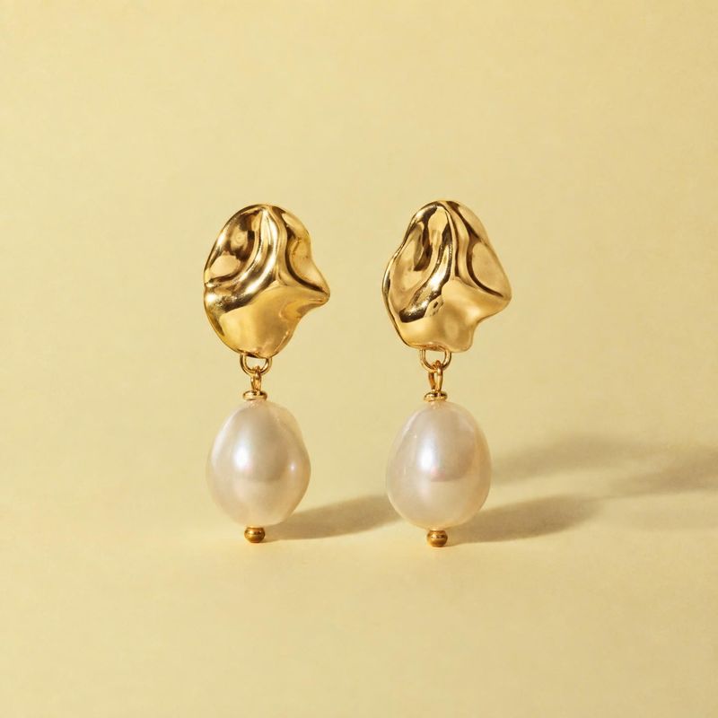gold and pearl drop earrings flat lay product photo: A pair of sculptural gold-tone earrings with luminous white pearl drops stands against a pale yellow studi…