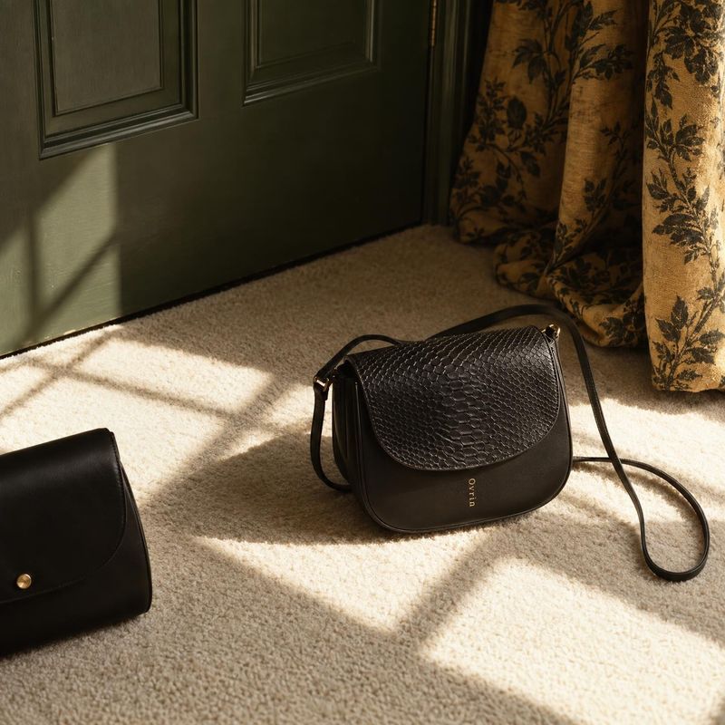 black shoulder bags lifestyle scene product photo: Two black shoulder bags are arranged on a sunlit cream carpet beside a dark green paneled door and patterned…