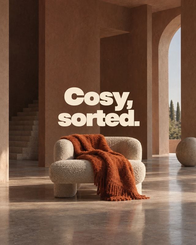 upholstered armchair room set product photo: A plush cream armchair draped with a rust-colored throw sits in a sculptural, warm-toned interior beneath bold cre…