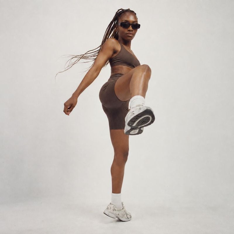 athletic outfit and sneakers action & motion product photo: A model in a dark brown athletic set and white sneakers lifts one leg toward the camera against a s…
