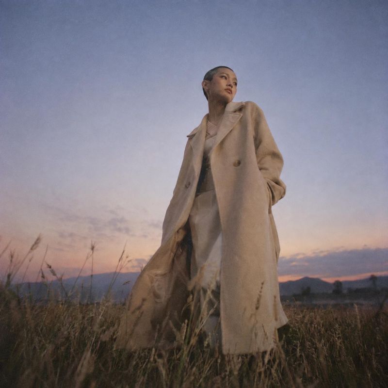 long beige coat autumn product photo: A model in a flowing beige coat stands in a grassy field at dusk, with a muted pastel sky and a quiet, cinematic mood.