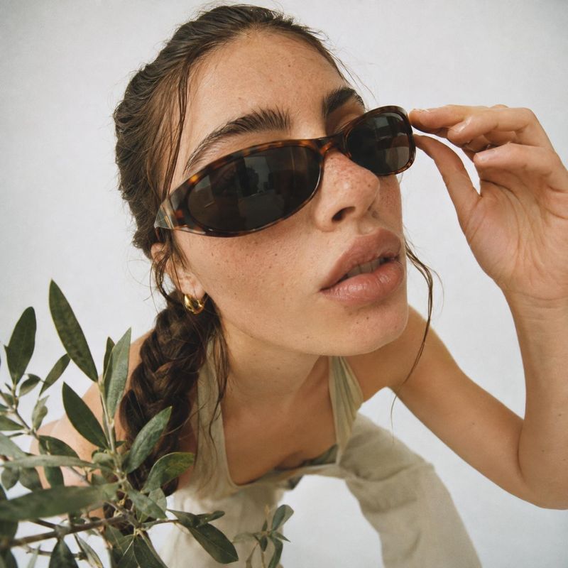 sunglasses on-body detail product photo: A woman in tortoiseshell sunglasses adjusts the frames in a close, softly lit studio portrait, with leafy greenery add…