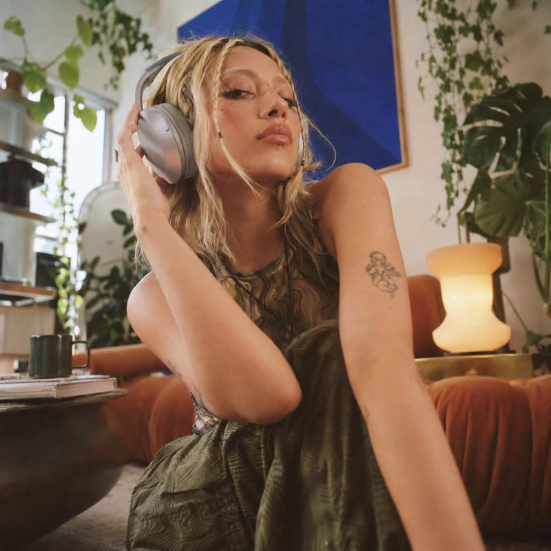 over-ear headphones equipment in use product photo: A woman in a patterned sleeveless outfit wears silver over-ear headphones in a plant-filled living room, ca…