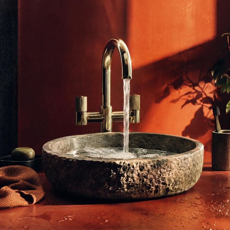 stone basin and brass faucet splash / pour product photo: A rustic stone basin and polished brass faucet fill a warmly lit terracotta bathroom, with running wa…