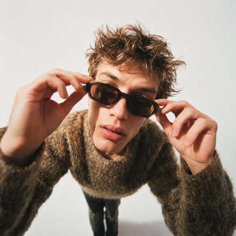 sunglasses on-body detail product photo: A young man in a textured brown sweater adjusts dark sunglasses in a close, low-angle studio portrait against a softly…