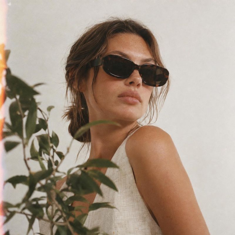 sunglasses on-body detail product photo: A woman in dark sunglasses and a light sleeveless top poses against a softly lit neutral wall, with leafy plants in th…