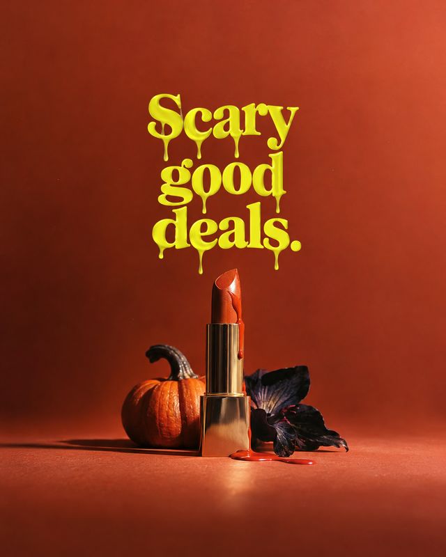lipstick halloween product photo: A red-orange lipstick stands beside a small pumpkin and dark foliage on a warm rust-colored studio set, with dramatic side li…