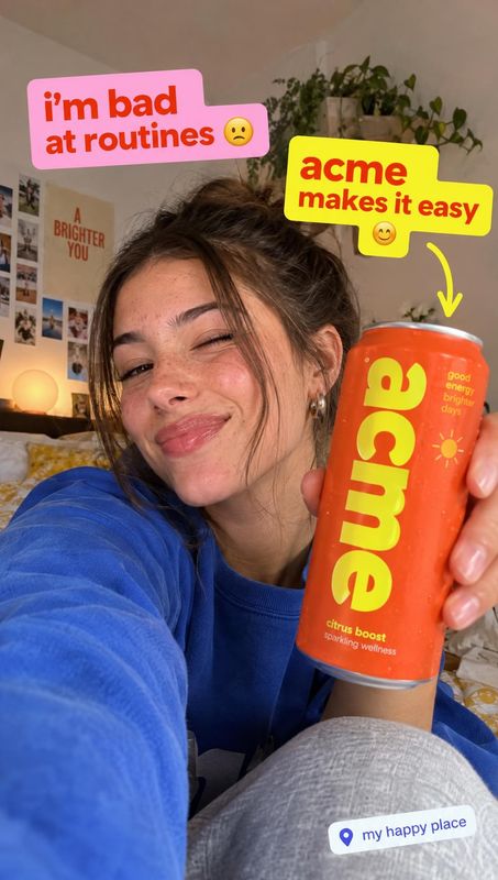 sparkling wellness drink lifestyle scene product photo: A woman in a blue sweatshirt smiles and holds an orange beverage can close to the camera in a cozy bedr…