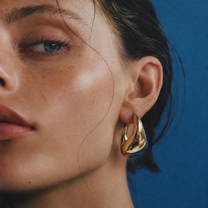 gold-tone statement earring on-body detail product photo: A close beauty portrait highlights a sculptural gold-tone earring against a vivid blue studio backdro…