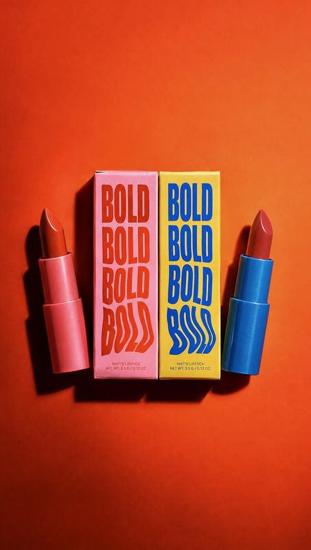 lipstick flat lay product photo: Two bold lipsticks and matching boxes are arranged on a vivid orange studio background, with contrasting pink and blue packagi…