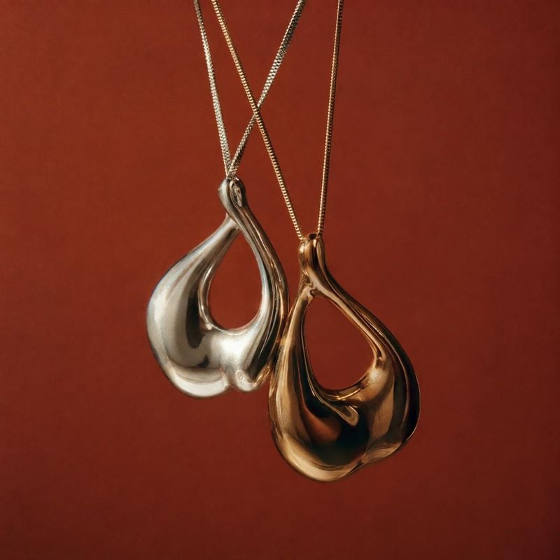 metal pendant necklaces close-up detail product photo: Two sculptural silver- and gold-tone pendant necklaces hang against a warm rust-red backdrop, lit with s…