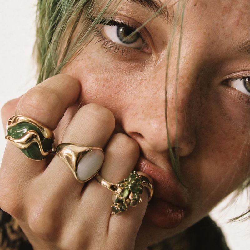 gold rings with gemstones close-up detail product photo: A tightly cropped portrait highlights a model wearing sculptural gold rings with green and pale stones…
