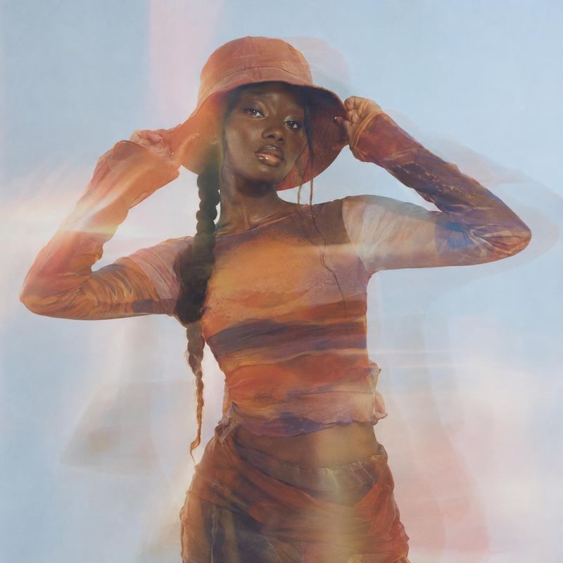 women's outfit and bucket hat action & motion product photo: A model in a sheer, warm-toned outfit and matching bucket hat poses against a pale blue studio bac…