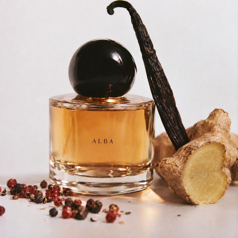 perfume ingredients product photo: An amber perfume bottle with a glossy black cap is styled among vanilla, ginger and scattered peppercorns on a pale studio s…