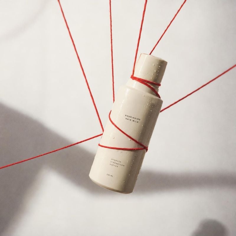 hair milk floating product product photo: A pale bottle of nourishing hair milk hangs at an angle, wrapped in taut red cords against a softly lit off-white stu…