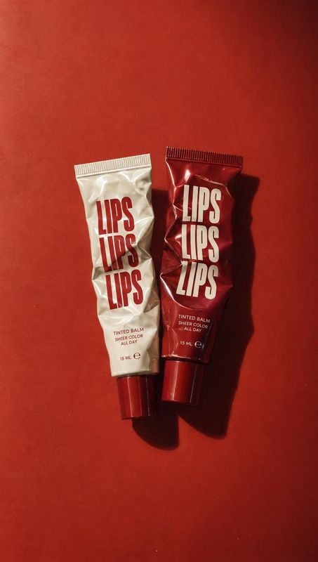 tinted lip balm flat lay product photo: Two crinkled lip balm tubes, one ivory and one deep red, rest on a vivid red surface in dramatic directional light with…