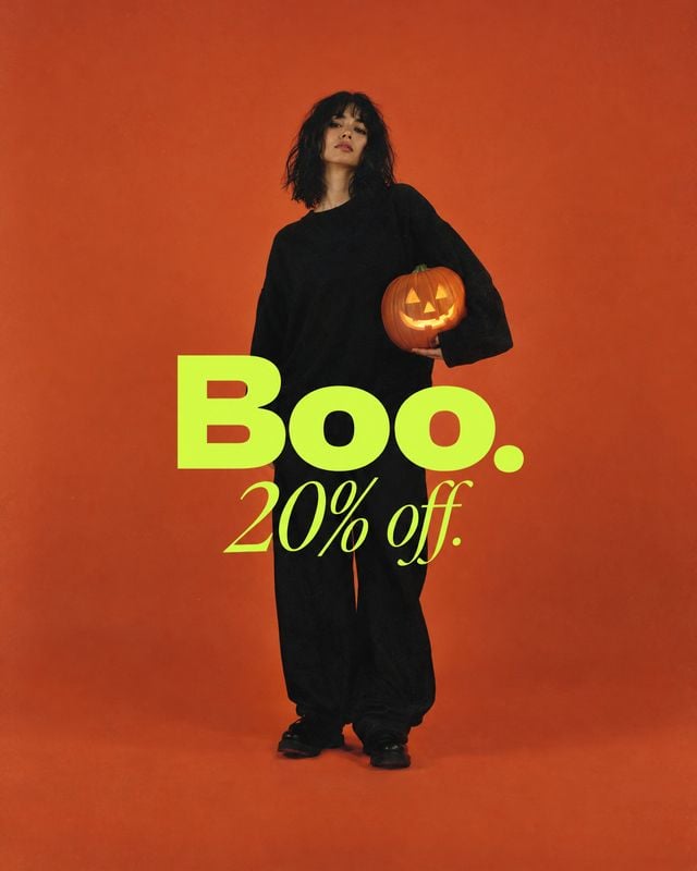 black outfit halloween product photo: A model in a loose black outfit holds a glowing jack-o'-lantern against an orange studio backdrop, with bold neon-green d…