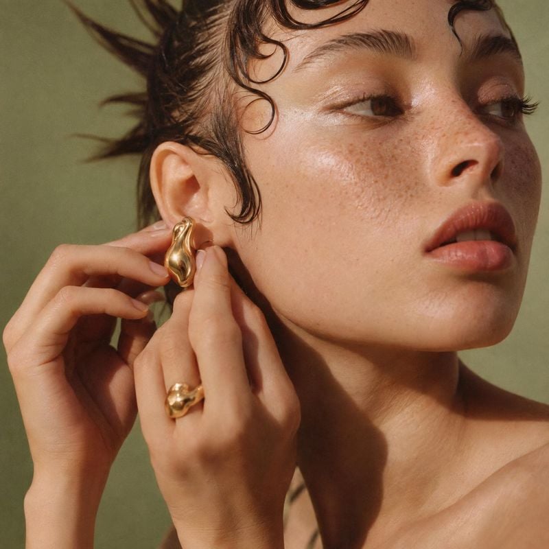 gold earrings and ring on-body detail product photo: A tight beauty portrait shows a woman fastening a sculptural gold earring, with a matching ring, dewy skin…