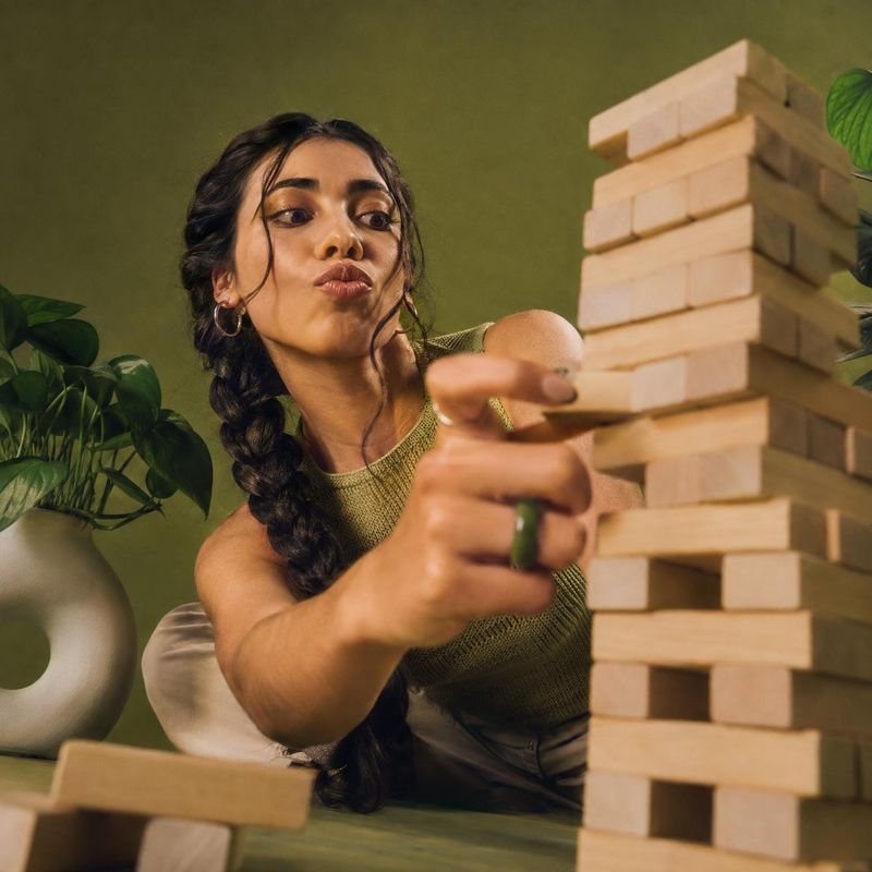 wooden stacking block game toys product photo: A woman in a textured green top reaches toward a tall wooden block tower against an olive-green studio set with…