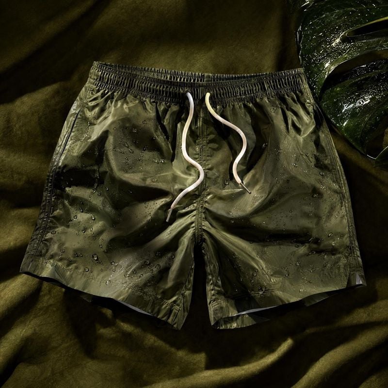 swim shorts flat lay product photo: Olive swim shorts with white drawstrings lie flat on dark green fabric, their wet sheen and water droplets picked out by dr…