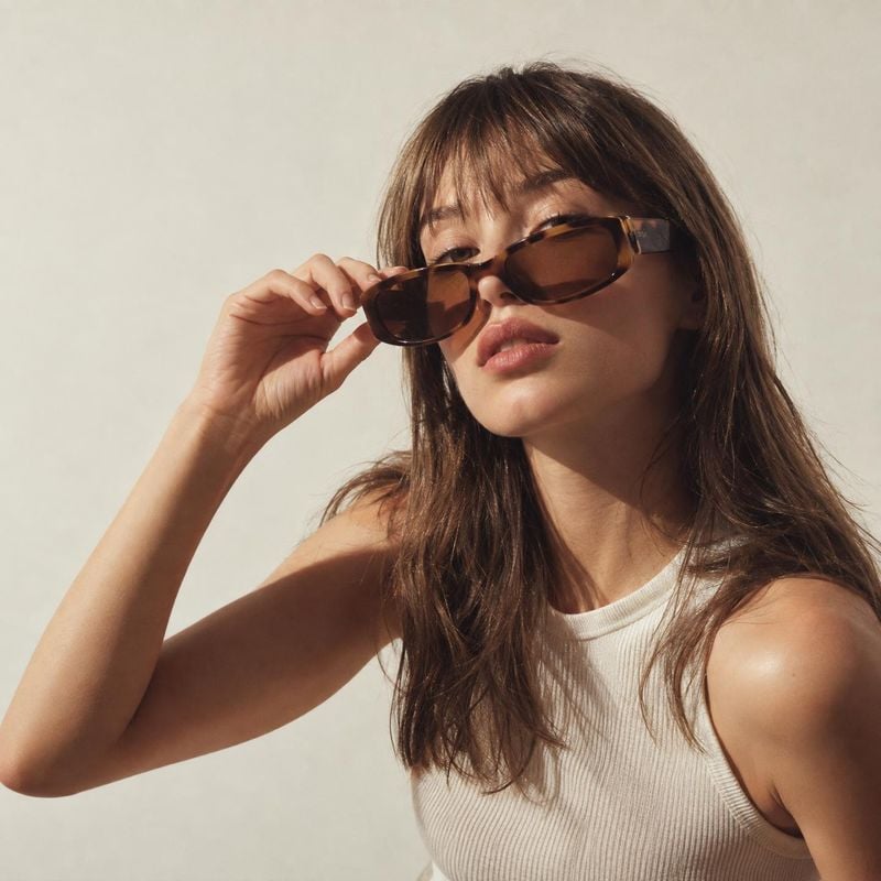 sunglasses on-body detail product photo: A woman in a white ribbed top adjusts oversized brown sunglasses against a warm, plain studio backdrop, with soft dire…
