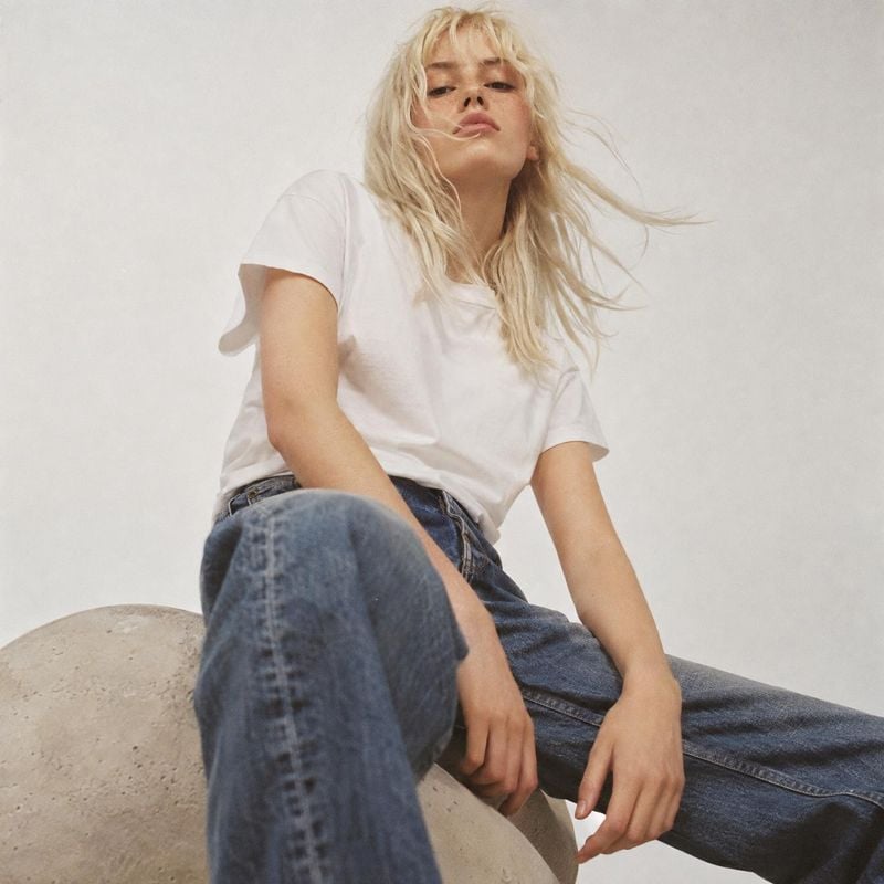 white T-shirt and blue jeans action & motion product photo: A blonde model poses in a white T-shirt and relaxed blue jeans against a pale studio backdrop, with…