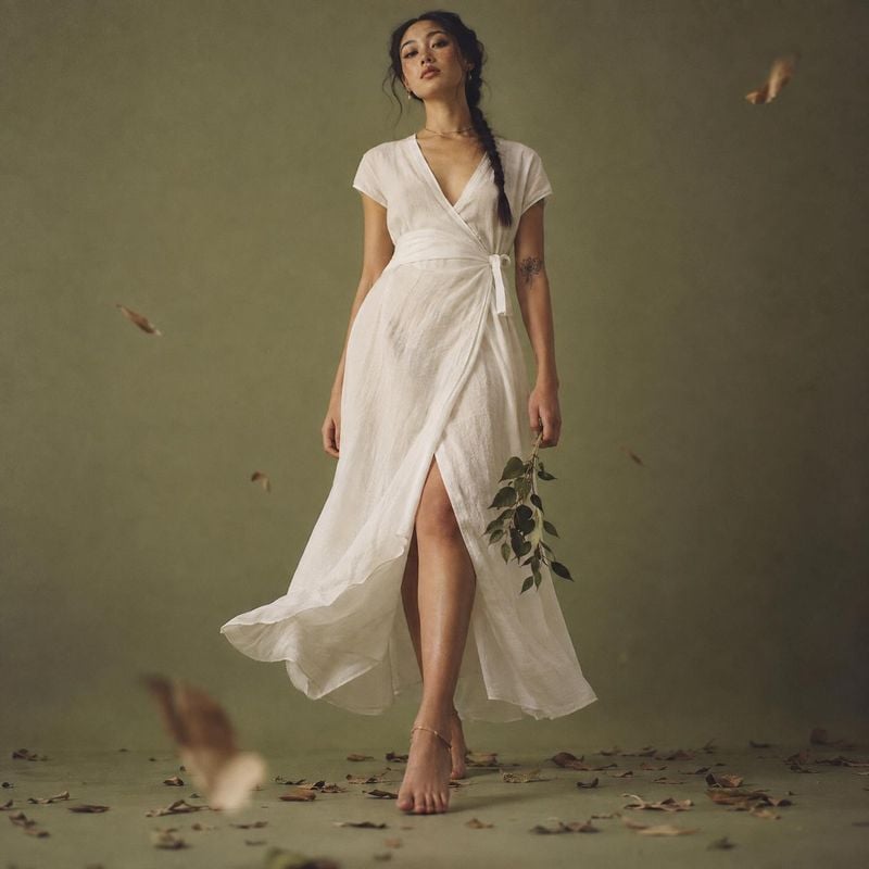 white wrap dress action & motion product photo: A model poses in a flowing white wrap dress against a muted olive studio backdrop, with scattered leaves and so…