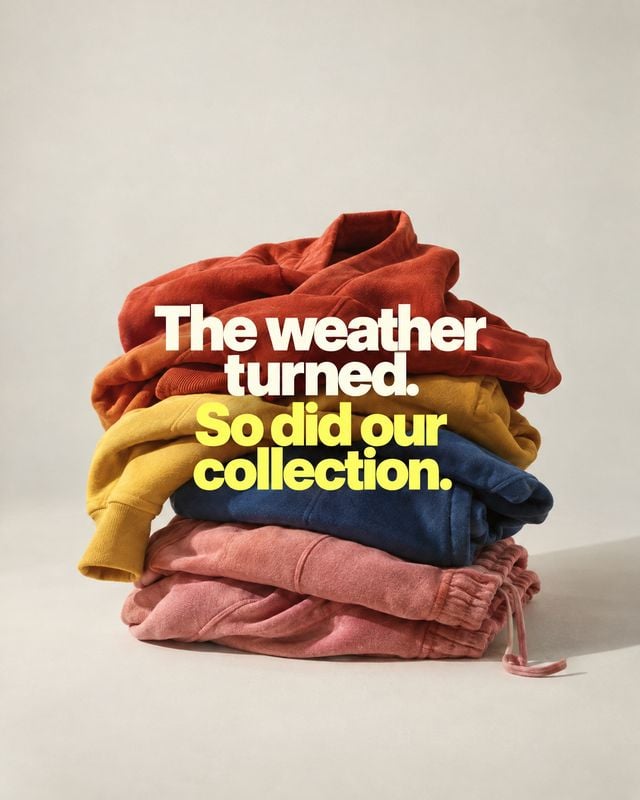 folded clothing collection group lineup product photo: A colorful stack of folded garments sits against a warm, neutral studio backdrop, with bold white and ye…