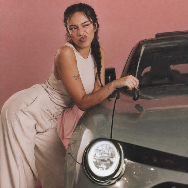 cream sleeveless outfit vehicle exterior product photo: A woman in a cream sleeveless outfit leans playfully against a dark car against a pink studio backdrop,…