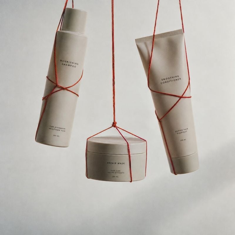 hair care set floating product product photo: A shampoo bottle, conditioner tube, and repair mask hang suspended by fine red cords against a softly lit neutral…