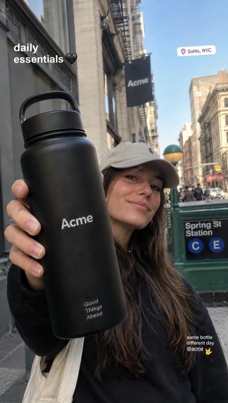 black reusable water bottle lifestyle scene product photo: A woman in a cap holds a matte black reusable bottle close to the camera on a sunny New York City st…