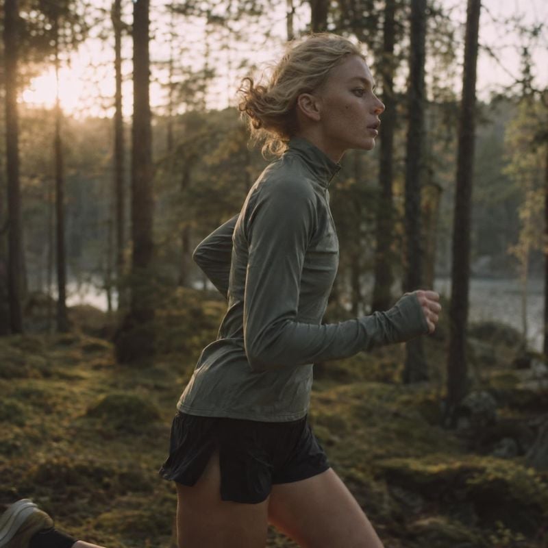 running apparel action & motion product photo: A woman runs through a forest in a muted gray athletic top and black shorts, backlit by warm low sunlight for a…