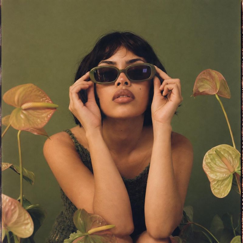 sunglasses on-body detail product photo: A woman in a textured dark top holds olive-toned sunglasses at her temples, framed by leafy plants against a muted gre…