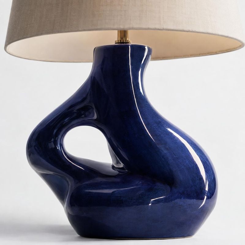 table lamp close-up detail product photo: A glossy sculptural navy-blue ceramic table lamp with a loop-shaped base and broad neutral fabric shade is photograph…