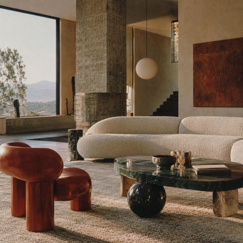 living room furniture interior space product photo: A sculptural living room with a curved cream sofa, richly colored lounge chairs, and a dark stone coffee ta…