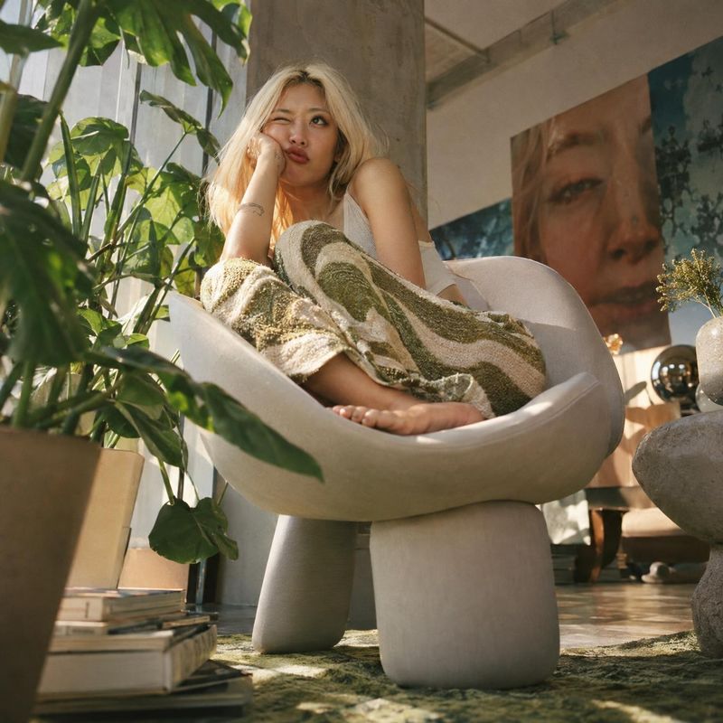 sculptural lounge chair lifestyle scene product photo: A blonde woman lounges barefoot in a sculptural cream chair, wrapped in a textured green-and-cream texti…