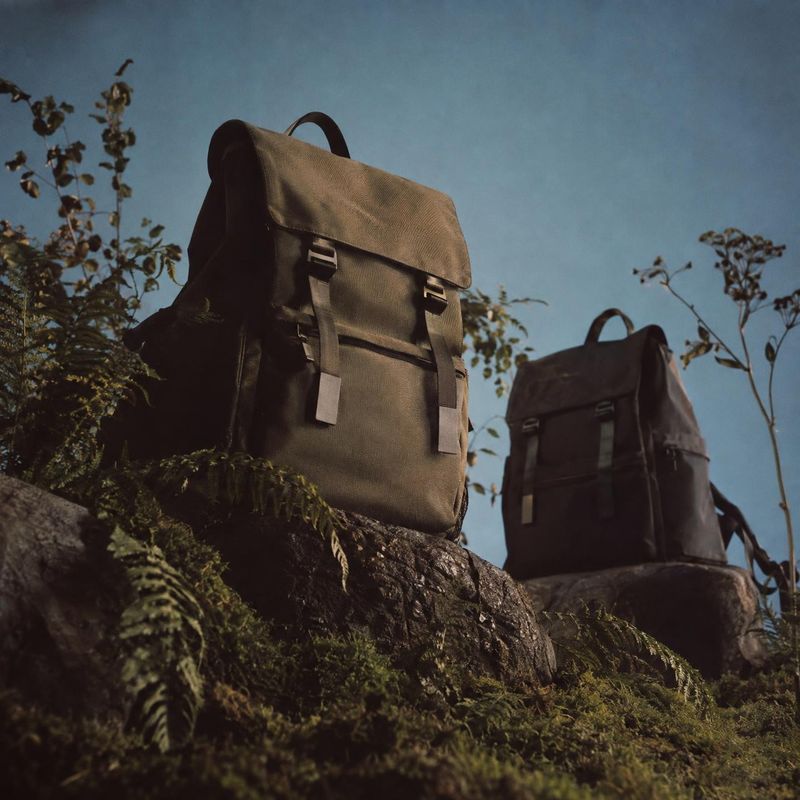 backpacks sports product photo: Two rugged canvas backpacks sit among rocks and ferns beneath a clear blue sky, lit with warm directional sunlight for an adven…