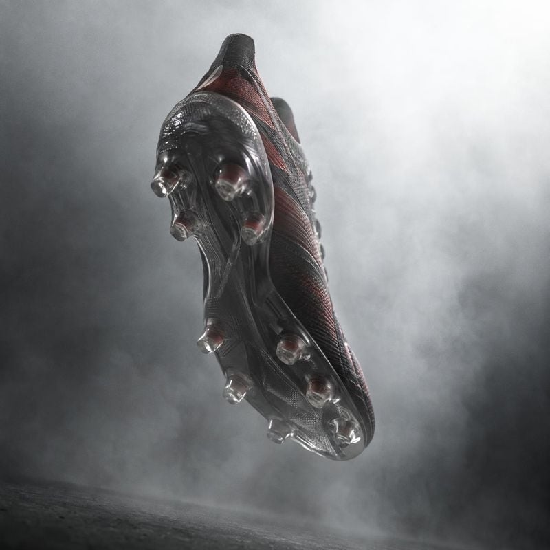 soccer cleat floating product product photo: A dark red-and-black soccer cleat floats against a smoky gray studio backdrop, lit dramatically to emphasize its t…