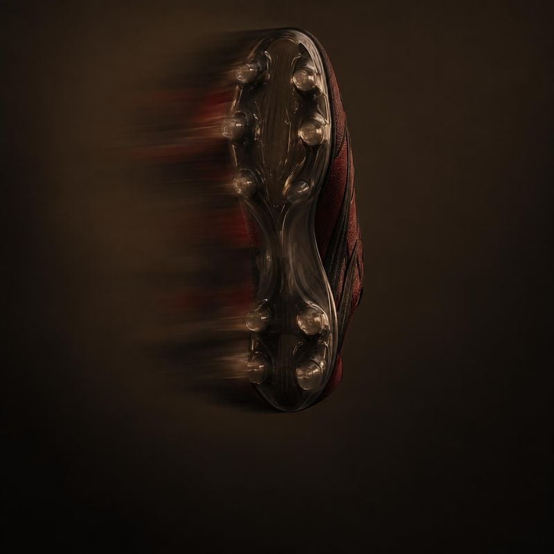 soccer cleat floating product product photo: A dark soccer cleat floats against a deep brown-black studio backdrop, its studded sole catching dramatic highligh…