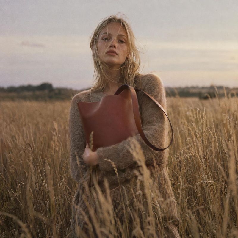 leather handbag autumn product photo: A woman in a soft neutral knit holds a structured brown leather handbag in a tawny field, photographed in warm, muted gol…