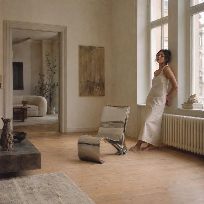sleeveless dress lifestyle scene product photo: A woman in a simple sleeveless ivory dress leans beside a tall window in a softly lit, neutral-toned interior w…