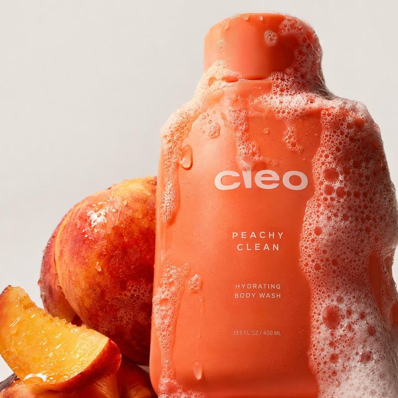 peach-scented body wash ingredients product photo: A peach-colored body wash bottle coated in suds is styled with fresh peach slices against a pale studio back…